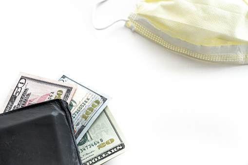 A twenty, fifty and hundred dollar bills fanned out and sticking out of a black leather wallet with a yellow face mask in opposite corner isolated on a white background with copy space.