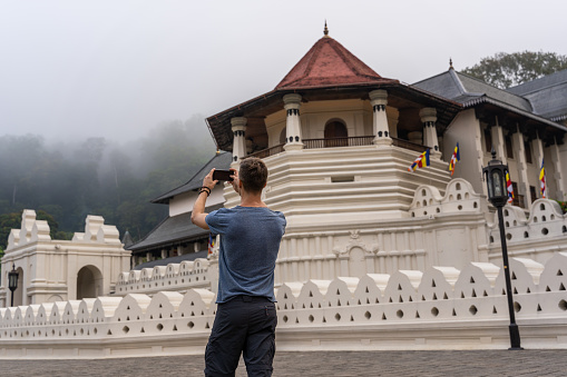 Caucasian boy use the smartphone to make picture of Temple of the tooth during foggy day in Kandy, Sri Lanka