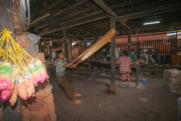 Mandalay Myanmar - November 5 2013; Confectionary factory with man pulling and stretching long string of toffee while women workers pack on bench in background.