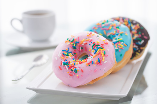 Pink, blue, and chocolate sprinkle donuts on a white plate and a cup of coffee on a light background.