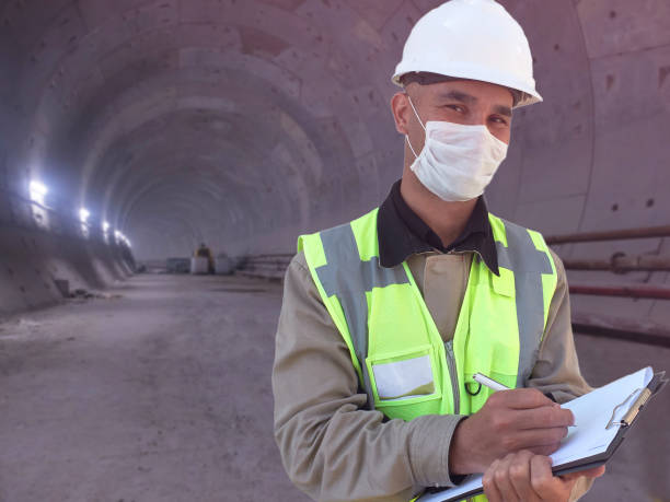 istock Construction worker dressed in medical mask, uniform and white hardhat stands with clipboard at metro tunnel 1225428592