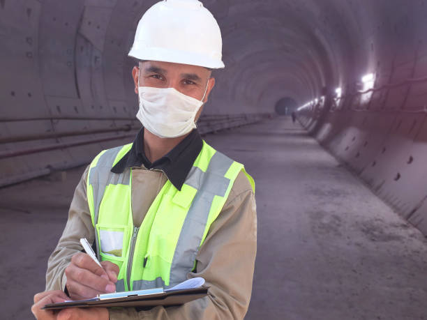 istock Construction worker dressed in medical mask, uniform and white hardhat stands with clipboard at metro tunnel 1225429795