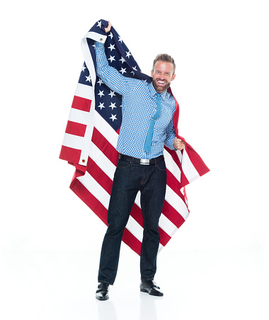 Full length of aged 30-39 years old caucasian male standing in front of white background wearing jeans who is successful and celebrating and showing patriotism and holding american flag