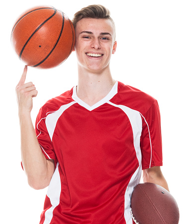 Front view of aged 16-17 years old caucasian young male american football player standing in front of white background wearing soccer uniform who is showing cool attitude and holding basketball - ball and playing soccer - sport and using sports ball