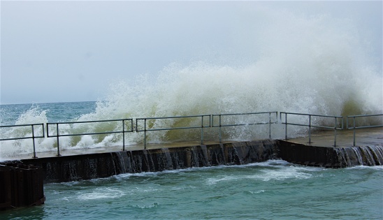 Overcast sky and waves crash against a railing, spilling over