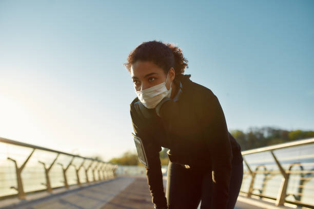 istock Exhausted african female jogger in protective mask resting after run while standing on the bridge in the morning. Jogging at sunrise. Sport and quarantine. Coronavirus 1225437890