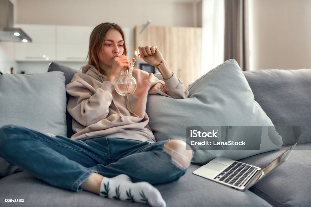 Need for Weed. Young caucasian woman sitting on the couch at home and lighting cannabis in the bowl of glass water pipe or bong. Cannabis and weed legalization concept Young caucasian woman sitting on the couch at home and lighting cannabis in the bowl of glass water pipe or bong. Cannabis and weed legalization concept. Horizontal shot Bong Stock Photo
