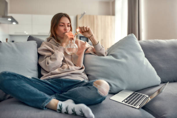 Need for Weed. Young caucasian woman sitting on the couch at home and lighting cannabis in the bowl of glass water pipe or bong. Cannabis and weed legalization concept Young caucasian woman sitting on the couch at home and lighting cannabis in the bowl of glass water pipe or bong. Cannabis and weed legalization concept. Horizontal shot bong photos stock pictures, royalty-free photos & images