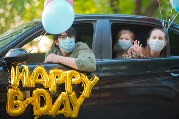 family wishing happy birthday to a friend or relative from their car during an infectious disease epidemic. they are keeping social distancing and wearing protective masks - illness mask pollution car imagens e fotografias de stock