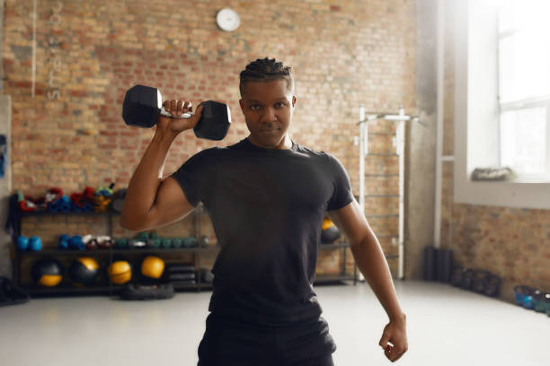 istock Hard Worker. Young athletic man looking at camera, lifting dumbbell while having workout at industrial gym. Sport, healthy lifestyle concept 1225446239