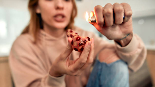 Necessary Tool. Young caucasian woman lighting cannabis in the bowl of glass pipe while sitting alone in the kitchen Young caucasian woman lighting cannabis in the bowl of glass pipe while sitting alone in the kitchen. Weed legalization concept. Selective focus. Horizontal shot pitter stock pictures, royalty-free photos & images