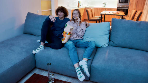Young teenage couple watching TV together, eating snacks after smoking marijuana from a bong or glass water pipe. They relaxing on the couch in the evening at home Young teenage couple watching TV together, eating snacks after smoking marijuana from a bong or glass water pipe. They relaxing on the couch in the evening at home. Cannabis, weed legalization bong photos stock pictures, royalty-free photos & images