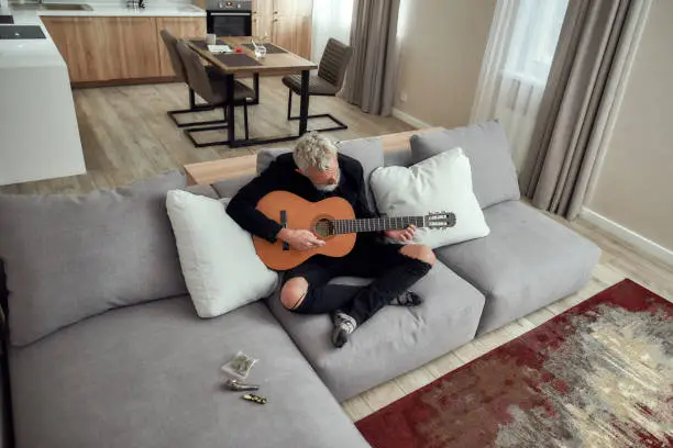 Photo of Outburst of the soul. Bearded middle-aged man, artist writing song, playing guitar at home. Metal pipe for smoking marijuana, cannabis buds in a plastic bag and lighter lying on the couch