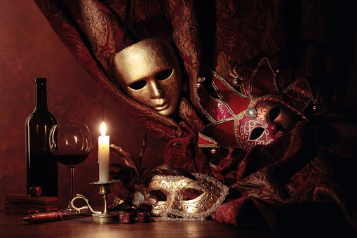 Carnival accessories and red wine on a old wooden table. Carnival masks and small brass candlestick with burning candle.