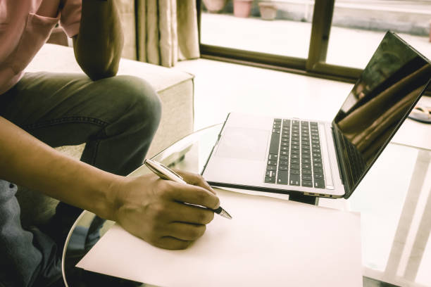 istock Anonymous Worker writing on a sheet of paper a business project work on office desk. Laptop is placed on the table. Side view. Close-up. Vintage style effect. Copy space room for text. 1225493958