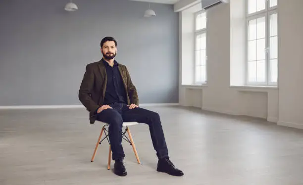 Full length of confident bearded man in stylish jacket looking at camera while sitting on chair in empty modern studio