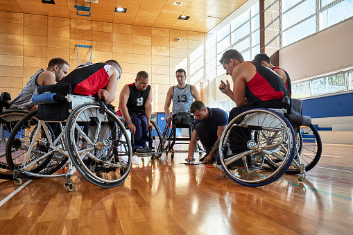 Low angle view of young and mid adult male wheelchair basketball players leaning-in to look at coach drawing play diagrams on clipboard.