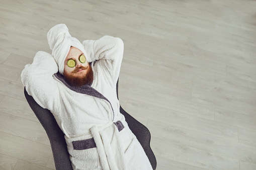 Funny weird fat bearded man with a cosmetic mask on his facein in bathrobe and a towel on his head makes spa treatments on his face resting against a gray background.
