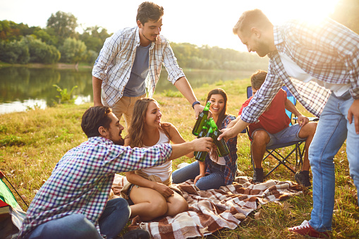 Young people drink, eat and clink glasses at a picnic in a forest near a lake in the summer in the autumn.