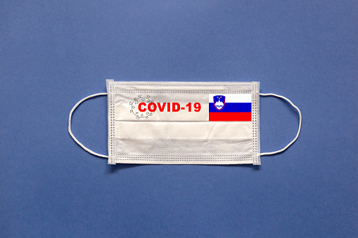 Slovenia flag and inscription COVID-19 on a medical mask on a blue background. Healthcare and medical concept. Pandemic virus COVID-19