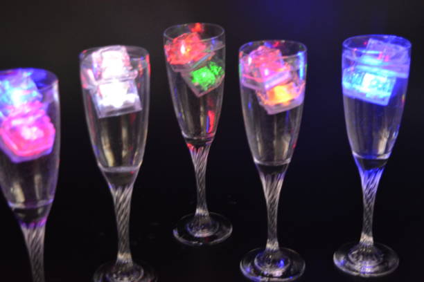 istock Five glass goblets on a long twisted legs with ice cubes glowing in different colors. Fun and bright drinks with luminous led elements are located on a black matte background, party and relaxation. 1225514441