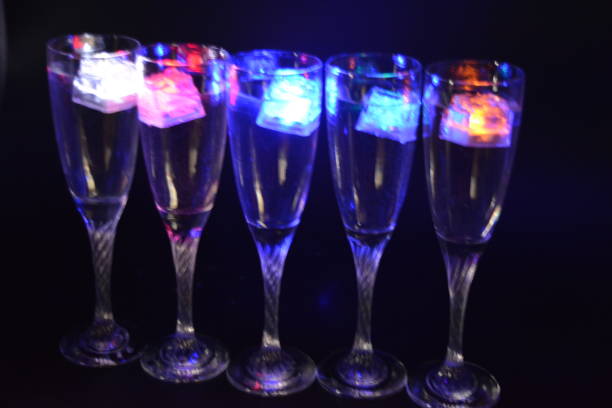 istock Five glass goblets on a long twisted legs with ice cubes glowing in different colors. Fun and bright drinks with luminous led elements are located on a black matte background, party and relaxation. 1225514502
