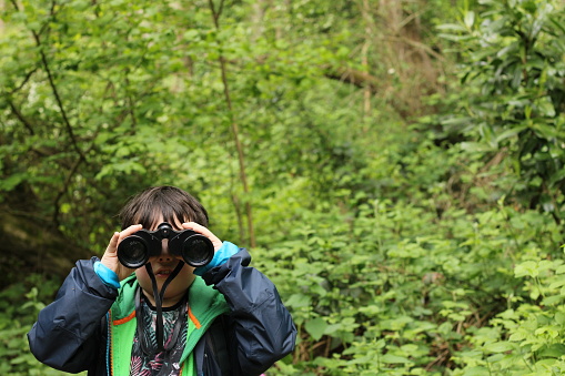 A child looking directly at the camera with binoculars on the left hand side of the picture the green forest background is blurred for copy space