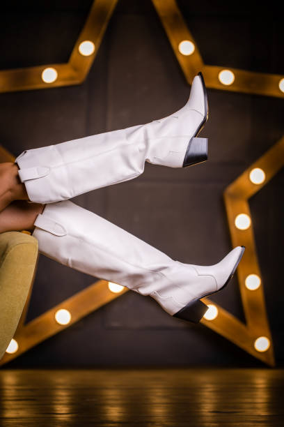 Boots below a white knee with a black heel dressed on a model that sits on a chair and grabs its legs, amid an LED star. Gray background with a gold star. studio shooting. thin legs. fashionable slippers.