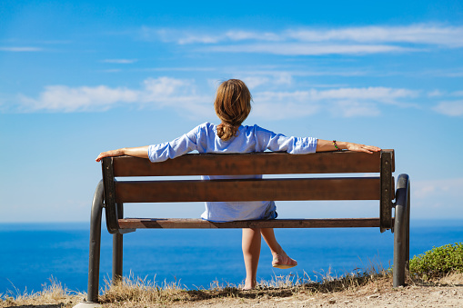 Woman sitting on a cliff bench above ocean / sea.