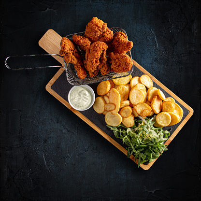 Flat lay of crispy fried chicken breast strips with golden potatoes and garlic sauce on dark background