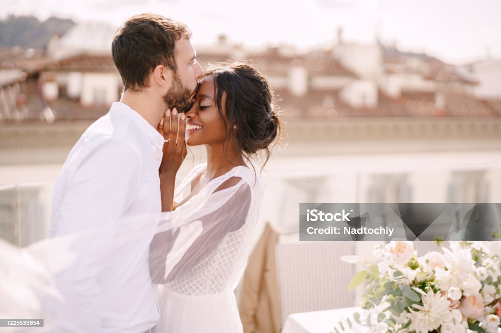 Destination fine-art wedding in Florence, Italy. Caucasian groom and African-American bride cuddling on a rooftop in sunset sunlight. Multiracial wedding couple Interracial wedding couple. Destination fine-art wedding in Florence, Italy. Caucasian groom and African-American bride cuddle on the roof, in the sunny sunset light. Wedding Stock Photo