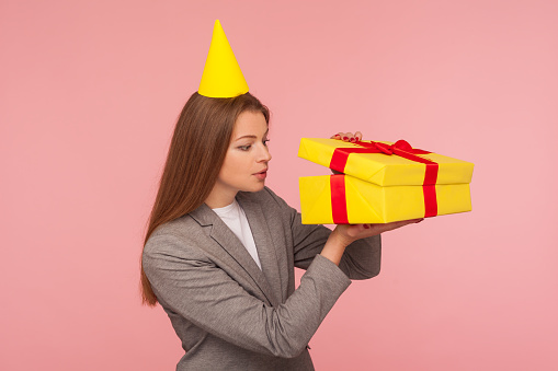 Portrait of nosy woman in business suit and with party cone on head looking inside present box, curious about gift, bonus, unwrapping birthday surprise. indoor studio shot isolated on pink background
