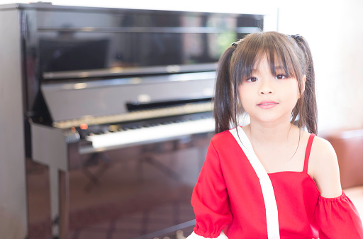 A portrait of a cute Asian elementary school girl wearing a red shirt sitting in the piano practice room