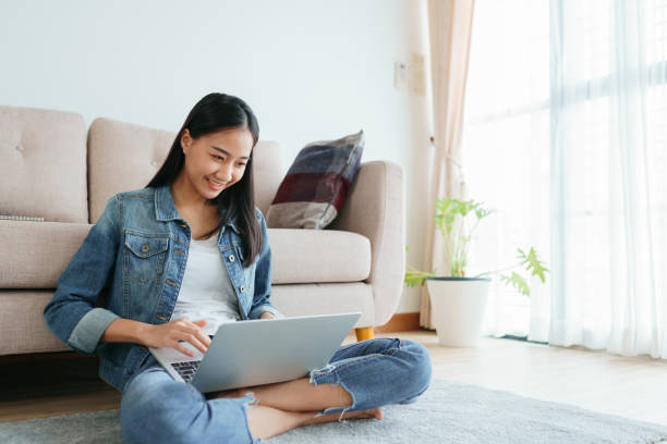 fille asiatique utilisant des jeans utilisant un ordinateur portatif tout en s’asseyant sur le plancher à la maison. les filles indépendantes font de la vidéoconférence avec leurs collègues sur les médias sociaux. concepts de travail de la maison e - collégienne photos et images de collection