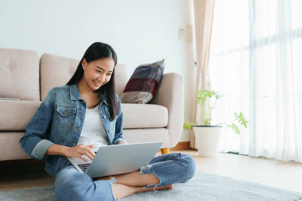 garota asiática usando jeans usando um laptop enquanto estava sentada no chão em casa. garotas autônomas estão em videoconferência com colegas nas redes sociais. conceitos funcionam de casa e novo normal - aluna da escola secundária - fotografias e filmes do acervo