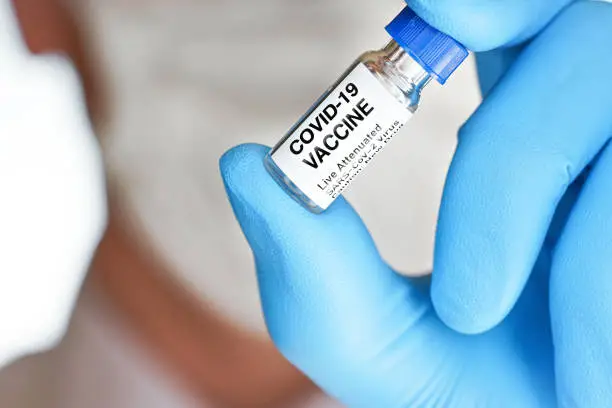 Hand in blue medical gloves holding small vial with label Covid 19 vaccine (sticker is own design, not real product), blurred face covered with cotton mask background. Coronavirus cure concept