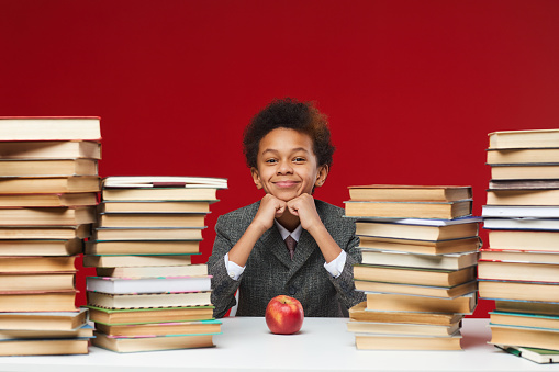 Front view portrait of cute African-American boy sitting at desk between stacks of books and smiling at camera, copy space