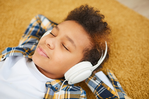Portrait of teenage African boy wearing headphones and lying on floor while enjoying music with eyes closed, copy space