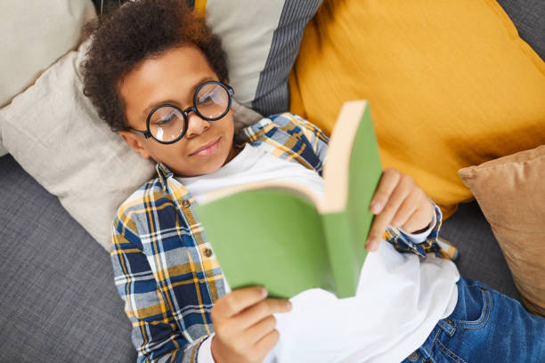Smart African Boy Reading Book Above view portrait of cute African boy wearing big glasses reading book while lying on couch, homeschooling concept, copy space reading stock pictures, royalty-free photos & images