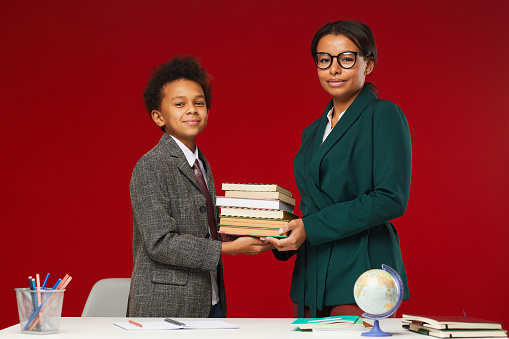 Portrait of young female teacher giving books to cute African-American boy in school, both looking at camera while standing against red background, copy space