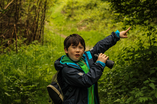 A picture of a young boy bird watching in the forest holding binoculars in one hand and excitedly pointing with the other green foliage background happy keen and excited face on the boy