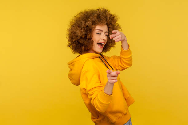 Hey you, handsome! Portrait of joyful curly-haired woman in urban style hoodie winking playfully Hey you, handsome! Portrait of joyful curly-haired woman in urban style hoodie winking playfully and pointing to camera, choosing guy and flirting. indoor studio shot isolated on yellow background blinking stock pictures, royalty-free photos & images