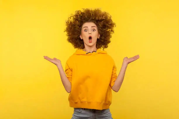 Photo of Portrait of extremely surprised girl in urban style hoodie jumping with her messy curly hair up, yellow background