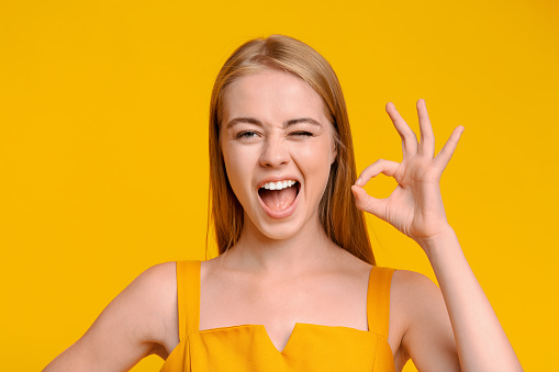 Everything is Great. Beautiful Young Girl Winking And Showing Ok Sign, Standing Over Yellow Background With Copy Space