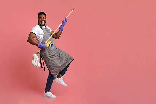 Funny African Man Using Mop Handle As Guitar, Having Fun During Cleaning. Posing Over Pink Background In Studio, Copy Space