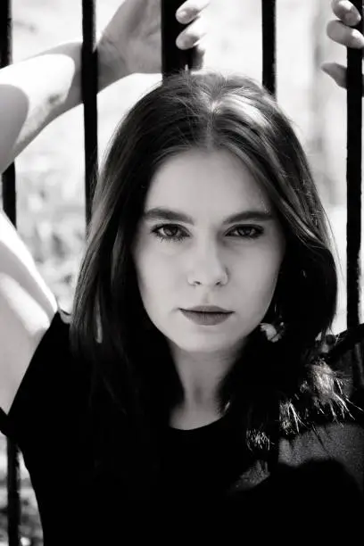 Black and white portrait of a beautiful young woman with long hair standing against an iron railing with arms raised staring intently at the camera with a thoughtful expression