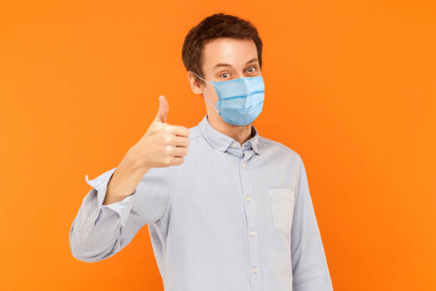 Like. Portrait of satisfied young worker man with surgical medical mask standing thumbs up and looking at camera smiling. Like. Portrait of satisfied young worker man with surgical medical mask standing thumbs up and looking at camera smiling. indoor studio shot isolated on orange background. ok sign photos stock pictures, royalty-free photos & images