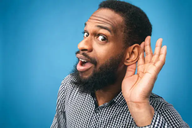 Eavesdropping Concept. Closeup of black guy holding hand near his ear and listening carefully isolated over blue wall