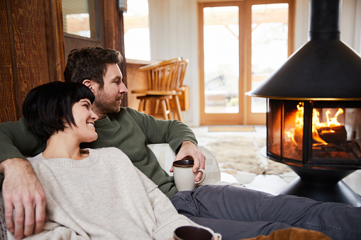 Smiling young couple relaxing on a sofa with cups of tea while warming up by their cabin fireplace