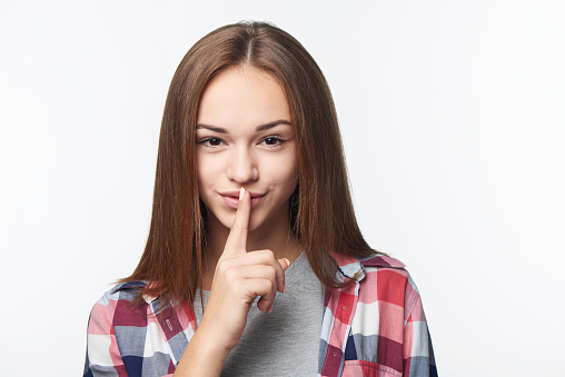 Close-up portrait of teen girl looking at camera showing finger on lips gesture, over light background
